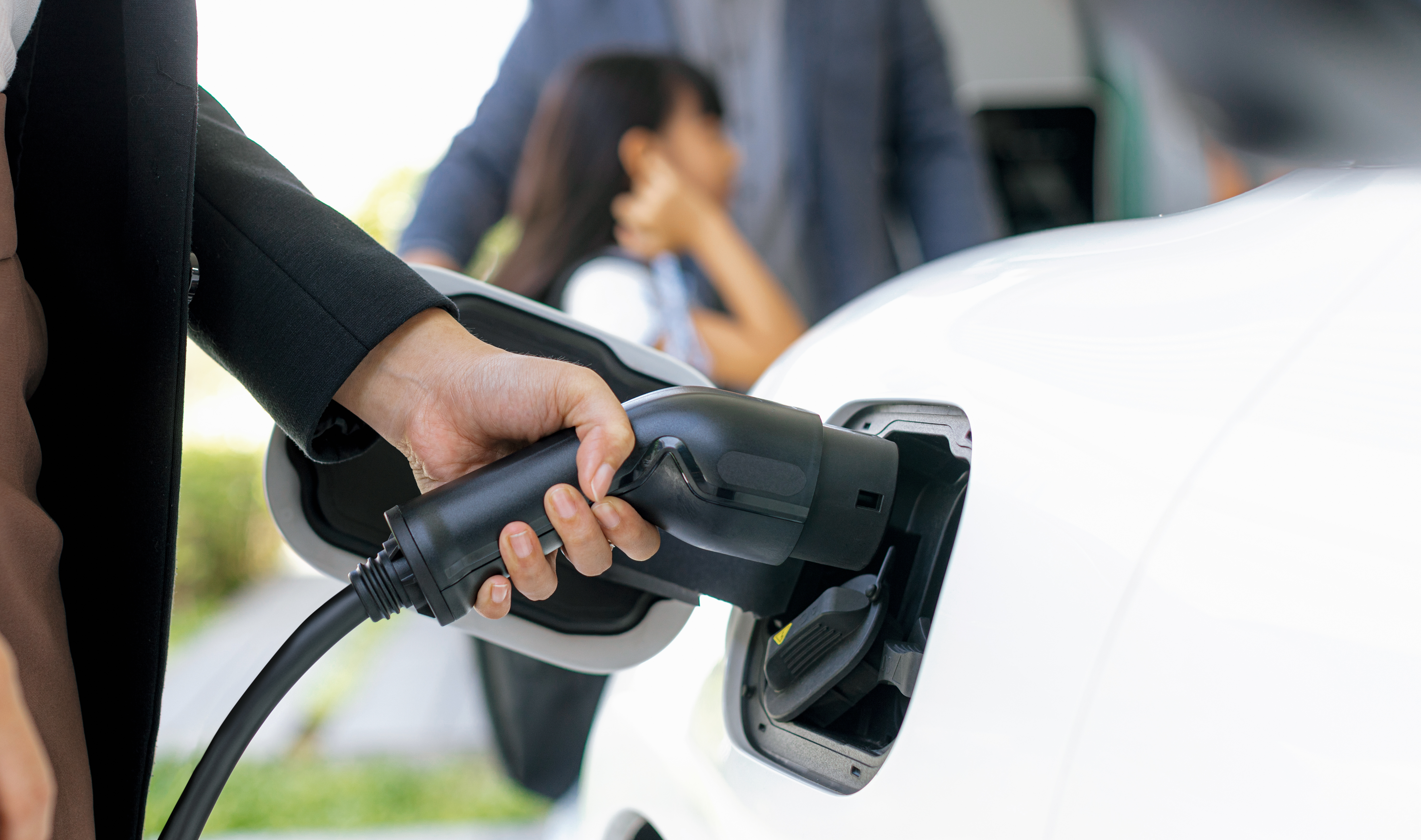 Focus closeup hand charging electric car, insert charger device into electric vehicle as progressive lifestyle concept of alternative green energy technology with blurred family in the background.