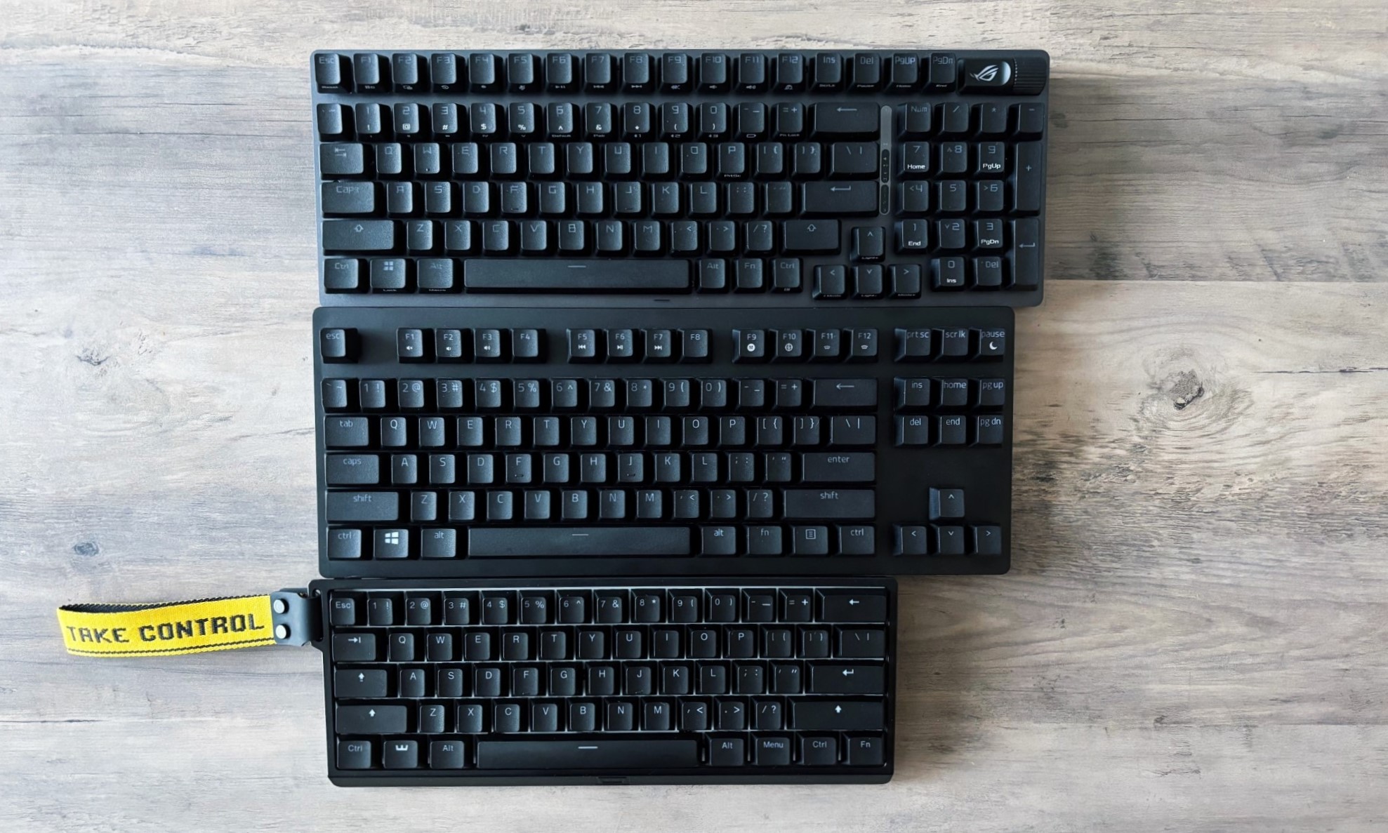 A trio of gaming keyboards of different sizes and layouts rest on a light brown wooden table. From top to bottom: A 96 percent keyboard, a tenkeyless (or 80 percent) keyboard and a 60 percent keyboard.