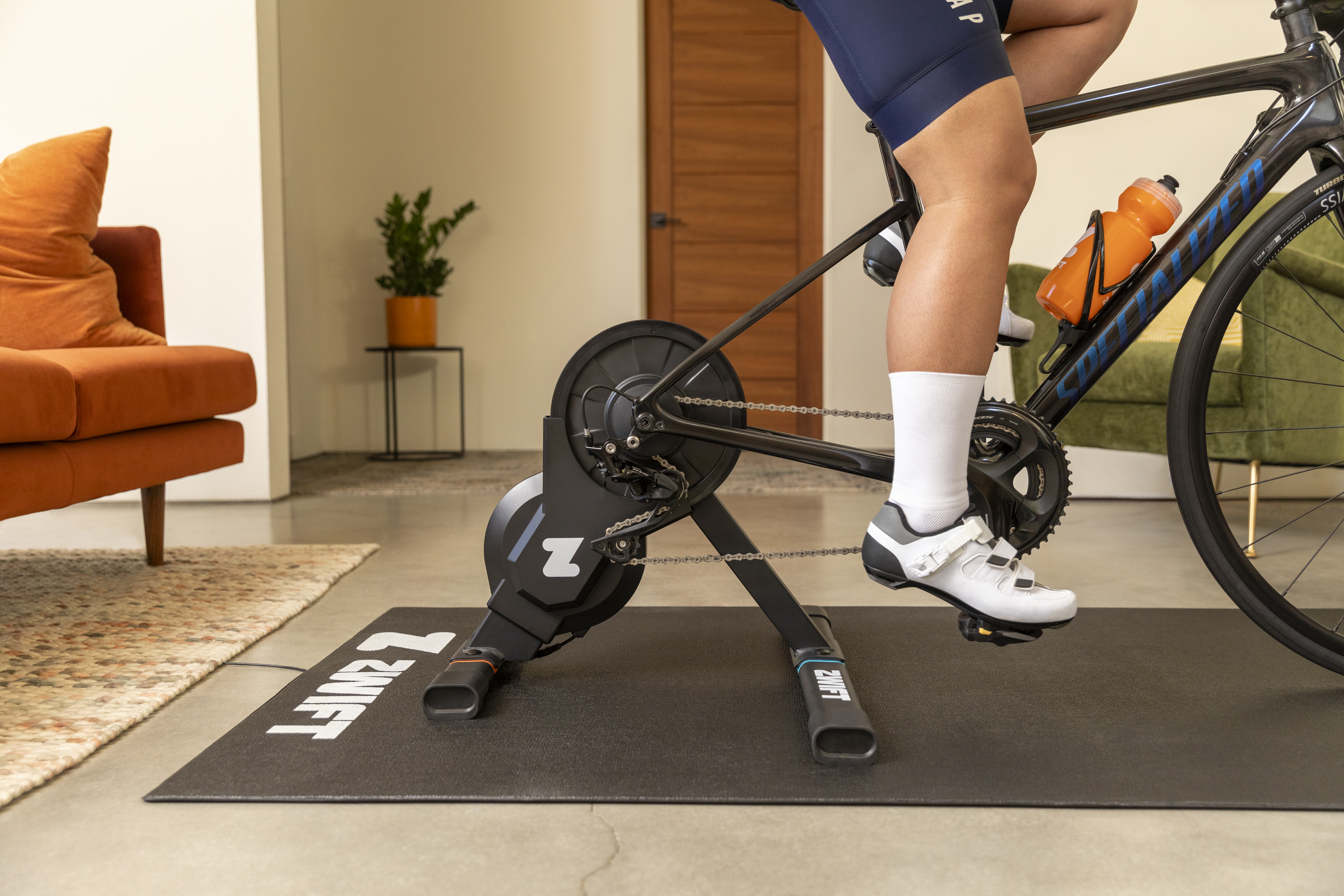 A sideview of the Zwift Hub One smart trainer in a living room with a bike connected and someone riding it.