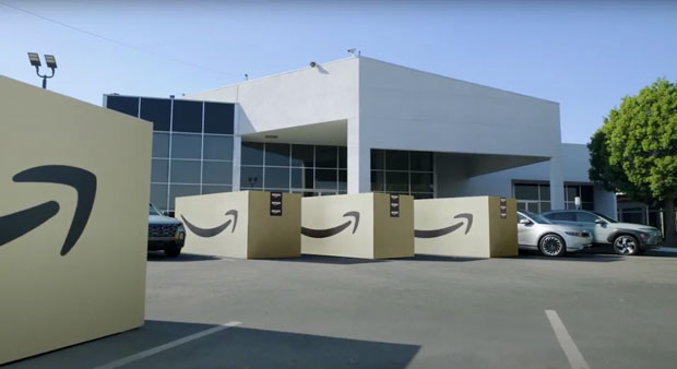 Promotional image featuring a white, featureless office block so favored by bland corporate types, in front of which are a series of car-sized Amazon packages.