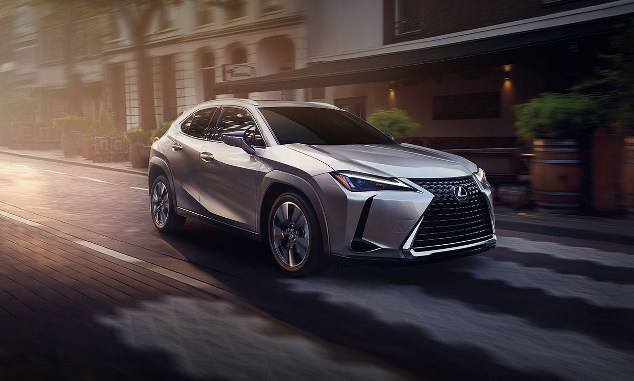 Foto di marketing del veicolo ibrido Lexus UX.  Il modello argento percorre una strada cittadina con un'illuminazione spettacolare.