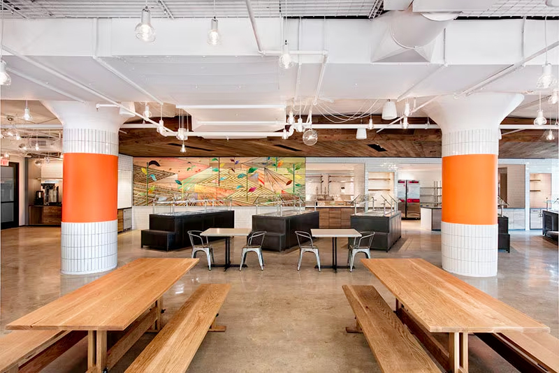 Etsy’s headquarters, inside view. A commons area includes benches and tables with pillars and buffet stations behind. Art decorates the walls.