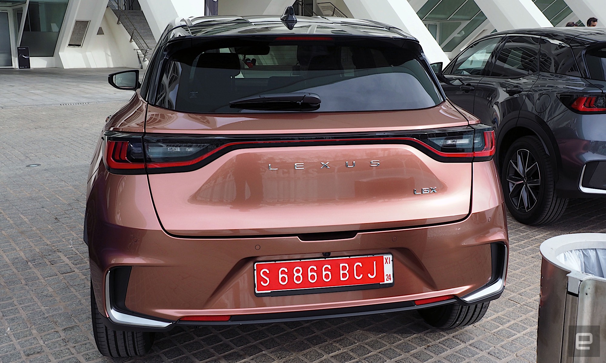 Image of a Lexus LBX trunk while parked under the canopy of the City of Arts and Sciences in Valencia. The car is painted in “Sonic Copper,” a sort of metallic orange. 