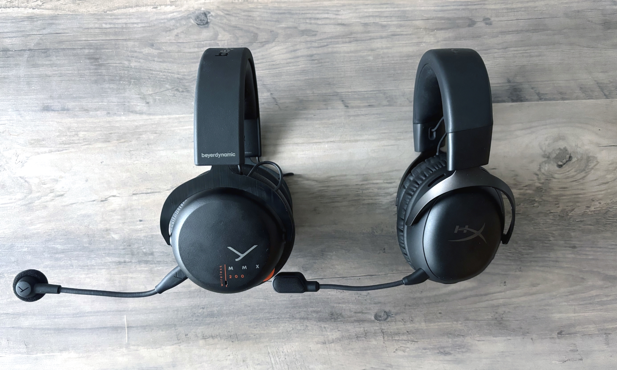 The Beyerdynamic MMX 200 and HyperX Cloud III Wireless gaming headsets lay on their sides on top of a brown wooden table.