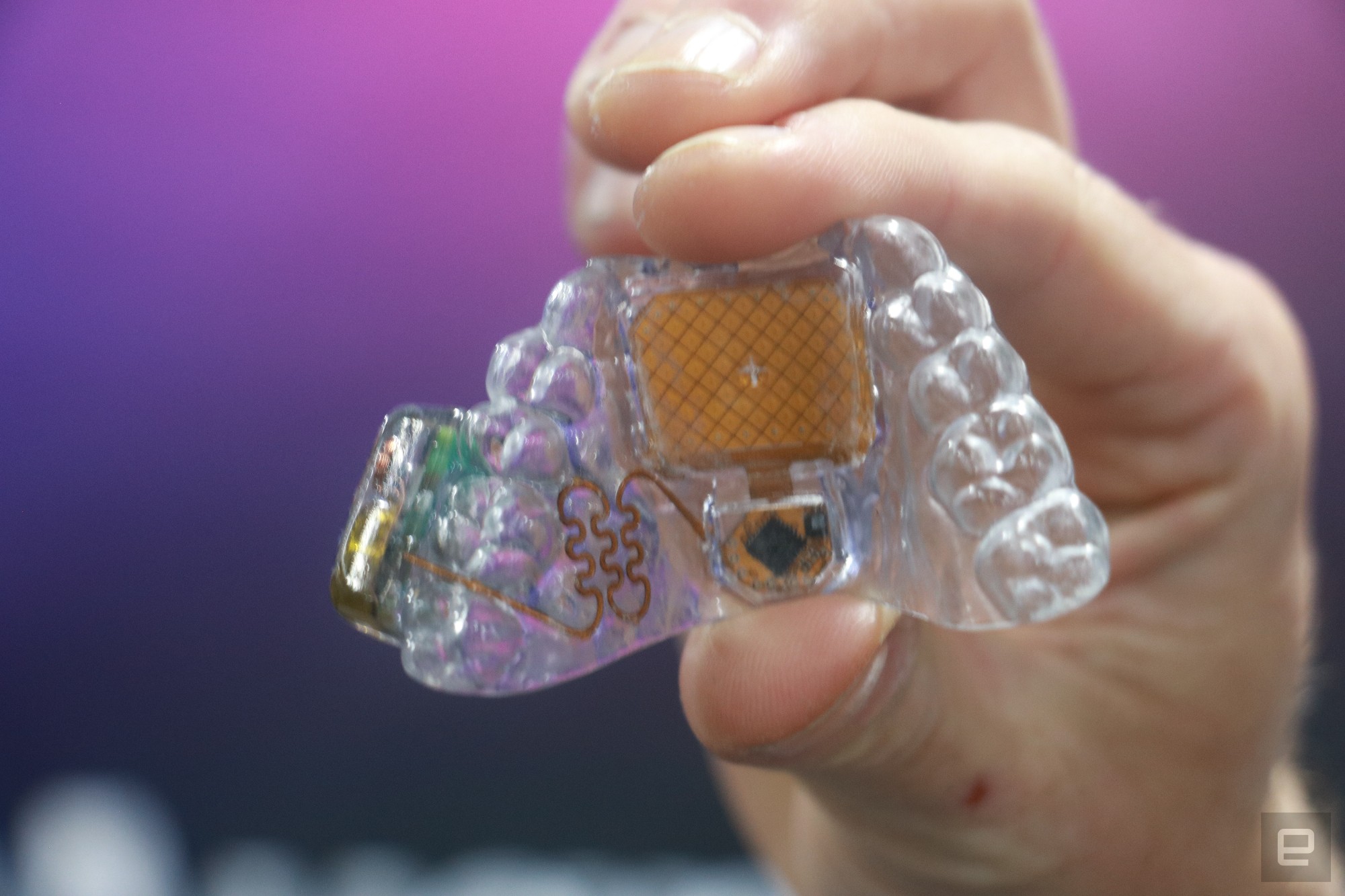 The MouthPad, a tongue-operate controller, held up in mid-air. It's a clear dental tray with an orange touchpad in the middle and some circuitry throughout.
