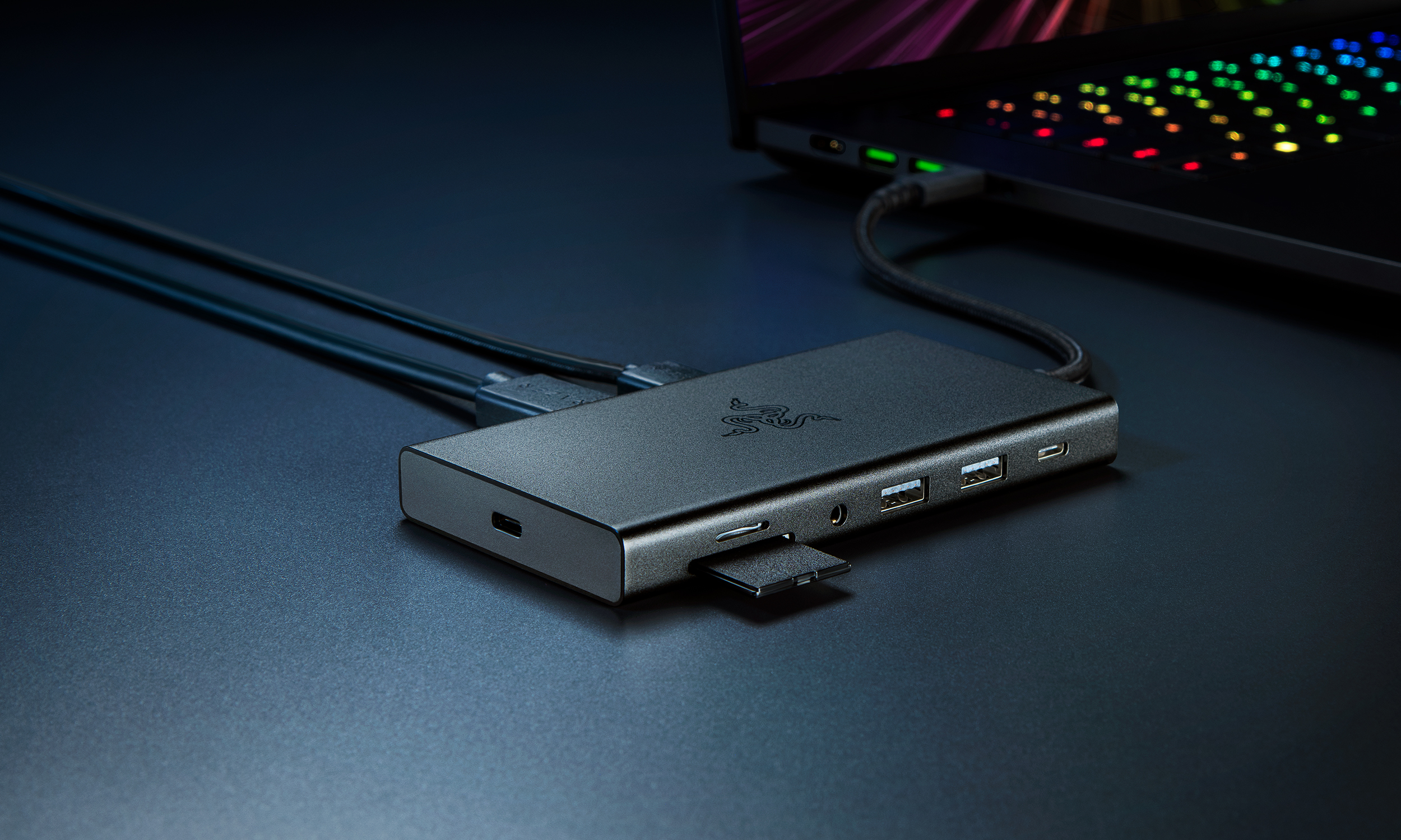 Product marketing photo of the Razer USB C Dock. The hub sits on a table with a gaming laptop behind it. It has several open ports and an SD card. It sits on a dark blue table with dramatic shades.