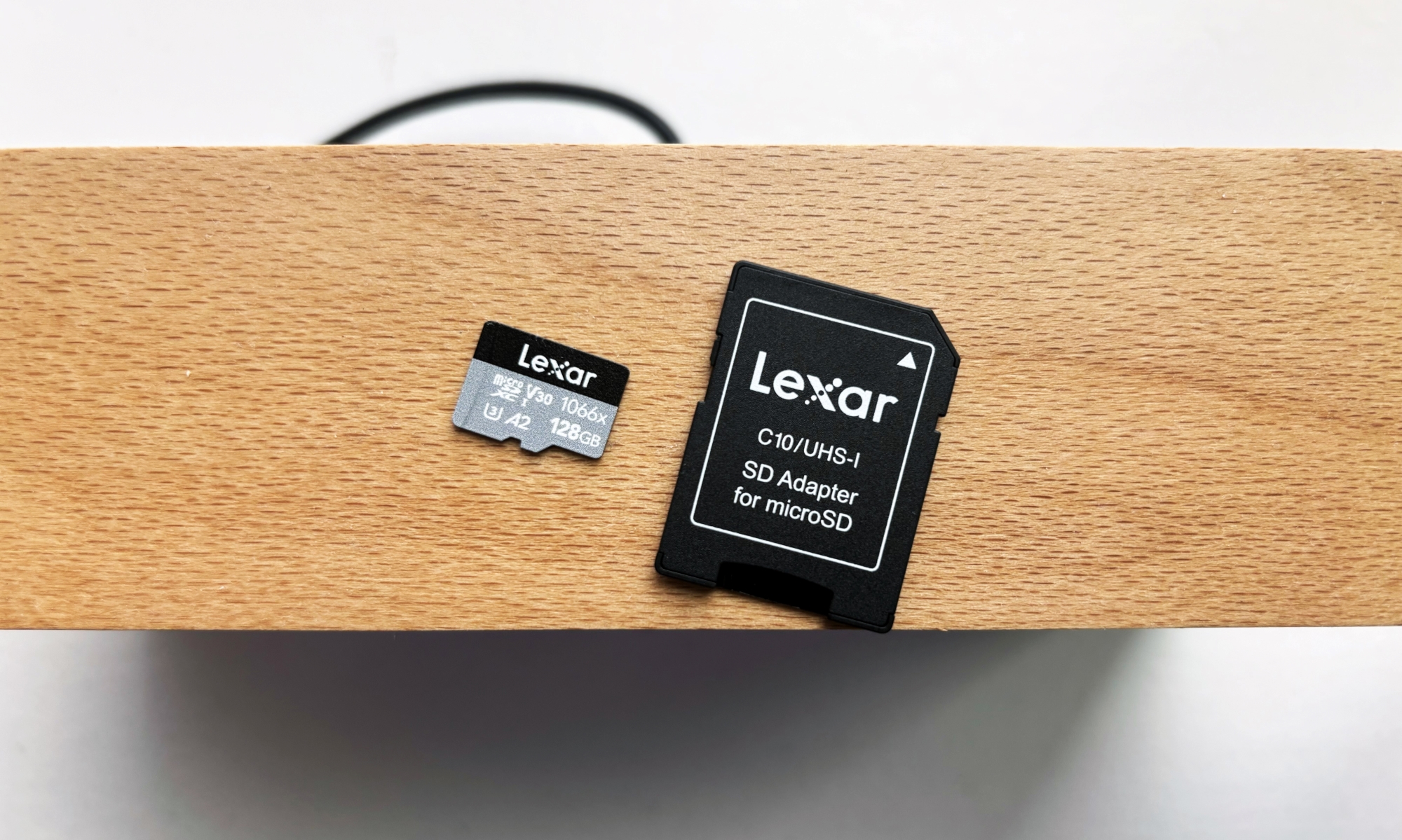 A silver and black Lexar Professional 1066x microSD card and its black SD card adapter rest on top of a brown wooden shelf above a white window sill.