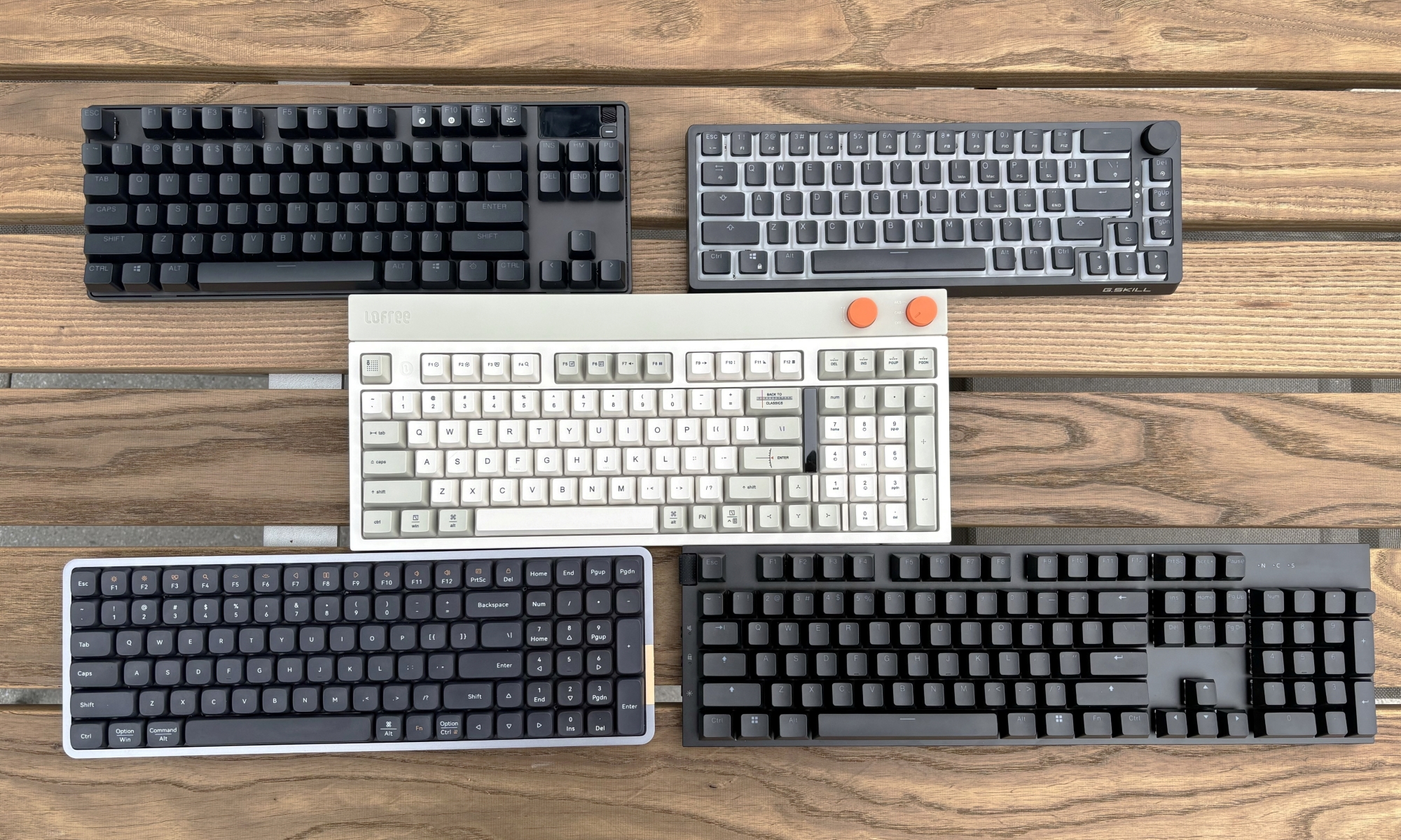 A quintet of mechanical keyboards rest on a brown wooden outdoor table, with one beige model flanked two separate keyboards above it, and two other models below it.