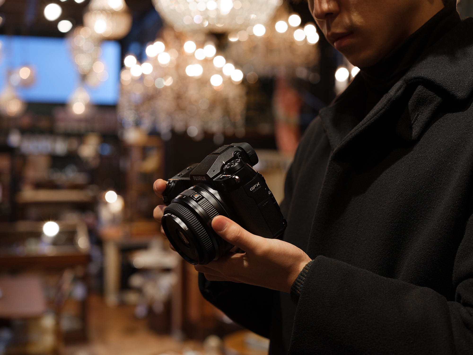 A person holding and looking down at the Fujifilm GFX 100S II in an indoor setting with blurred background. View from the person’s left side.