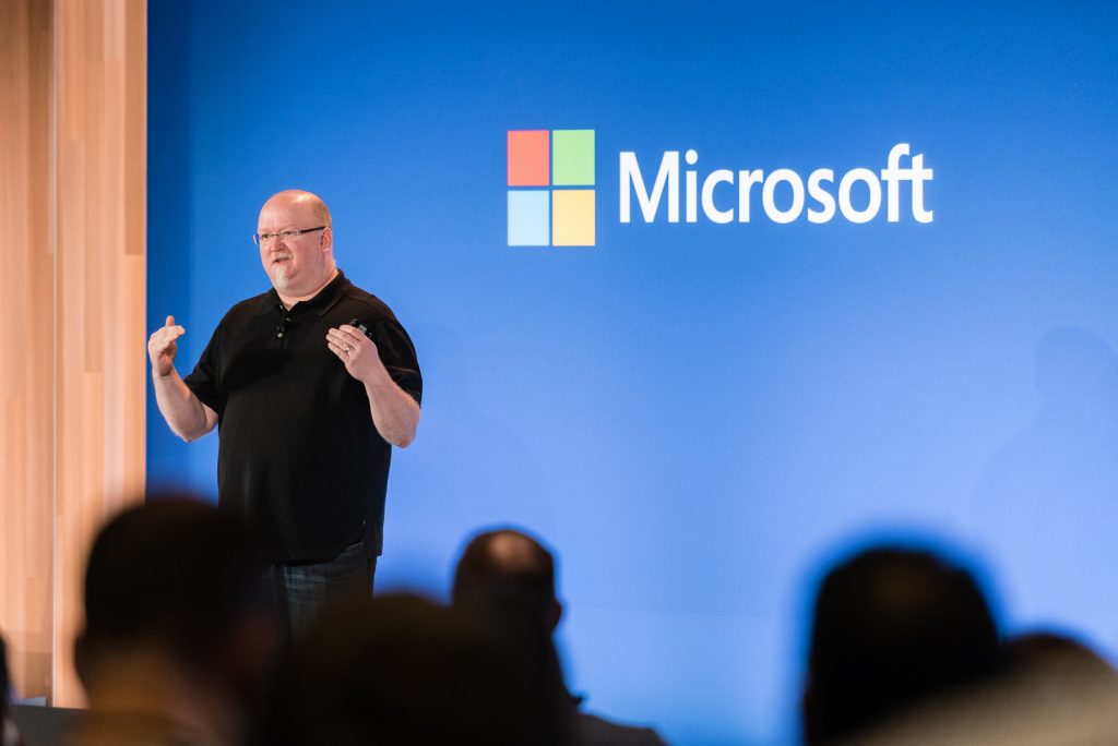 Microsoft CTO Kevin Scott presenting onstage in front of a blue wall with a Microsoft logo on it. Blurred audience heads in the foreground.