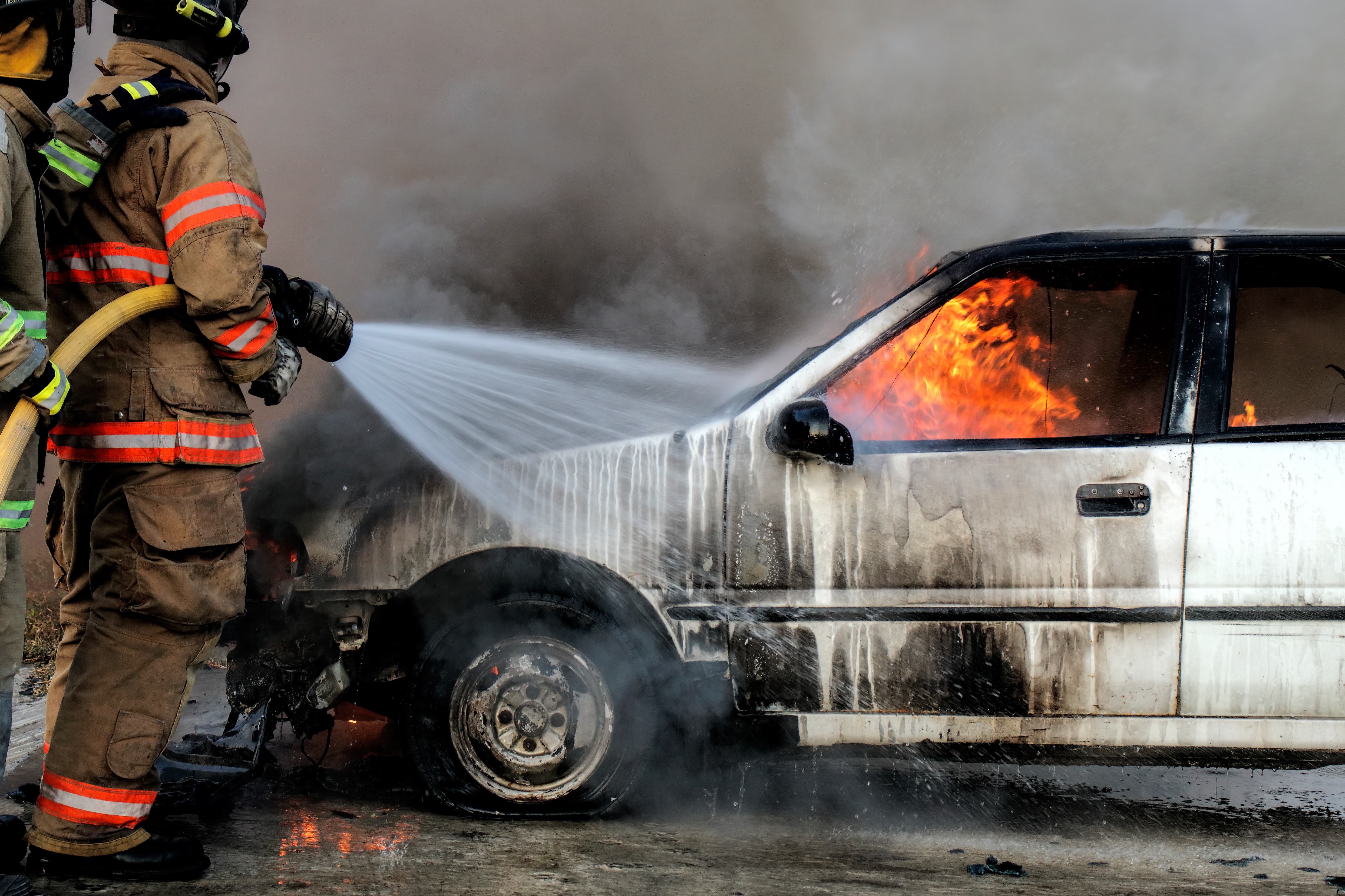 Firefighter in fire fighting suit spraying water, Firemen fighting raging fire with huge flames of burning car, Fire prevention and extinguishing concept