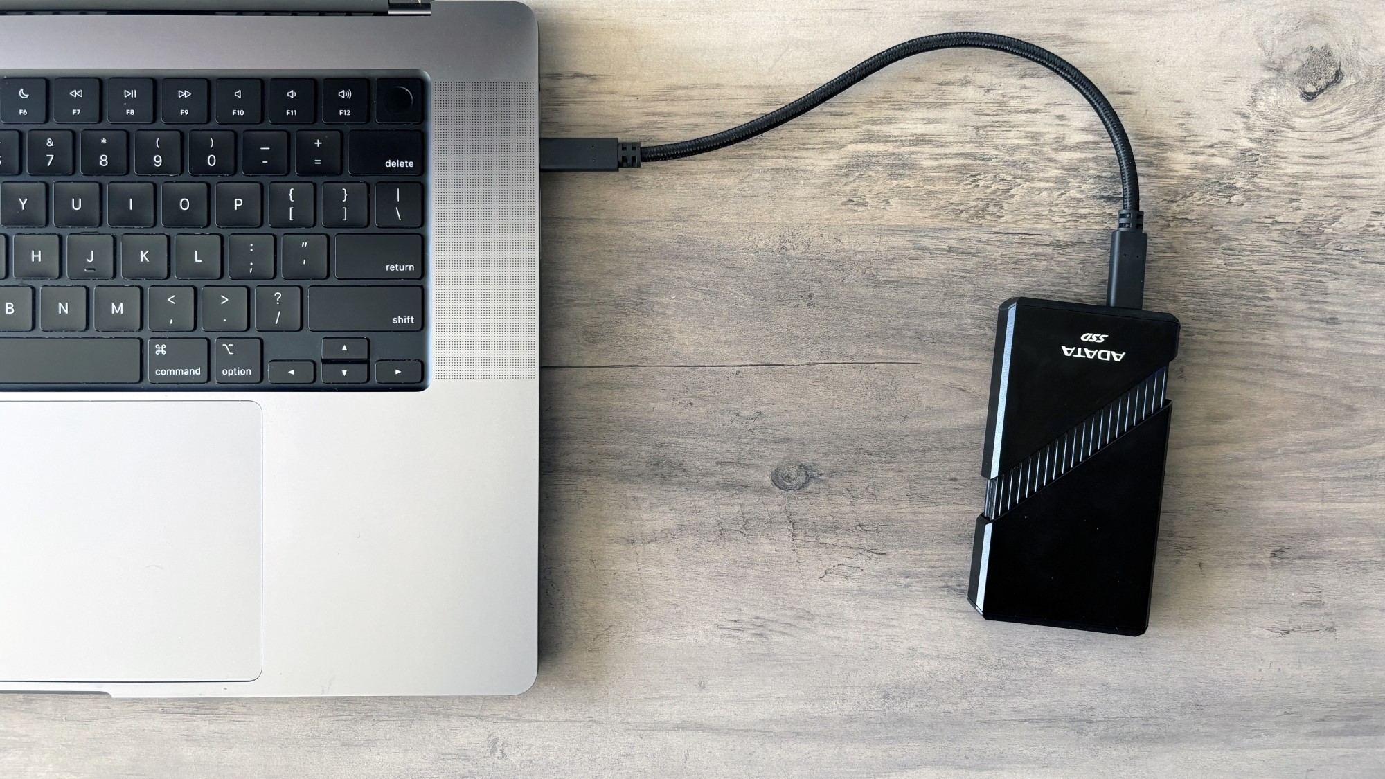 A portable SSD plugs into the side of a MacBook Pro on a brown wooden table.