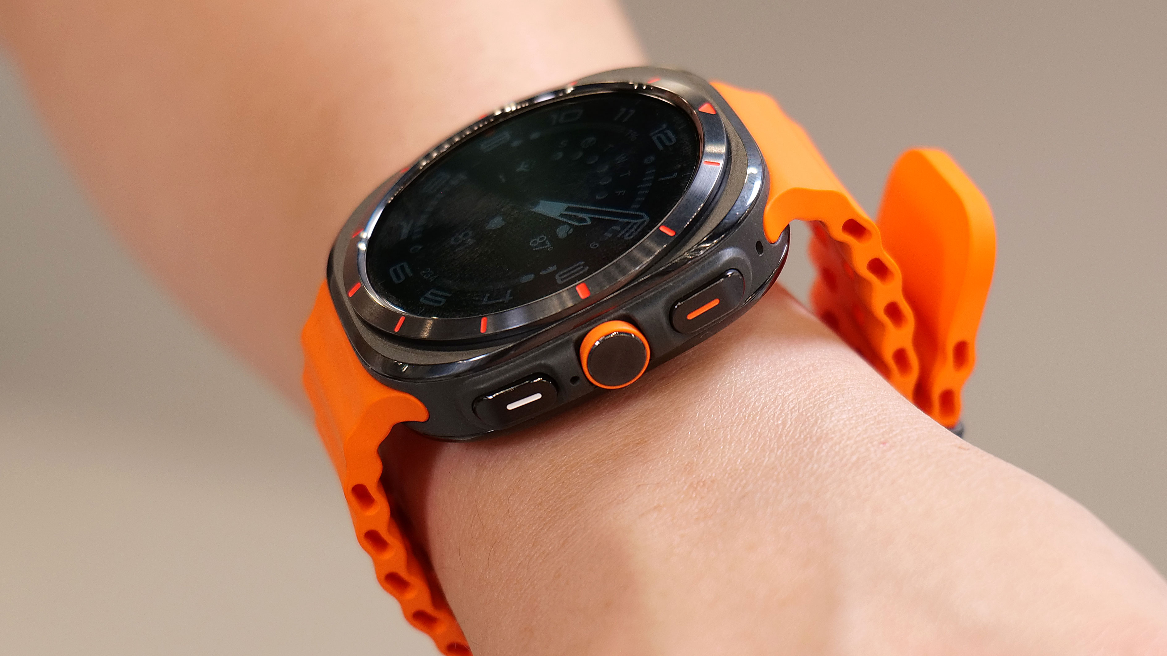 Side view of the Samsung Galaxy Watch Ultra on a wrist in mid air, showing the three buttons on its right edge. The top is a black rectangular button with an orange line in it, while the middle is a round dial with an orange outline. The last one is a black rectangle with a white line in it.