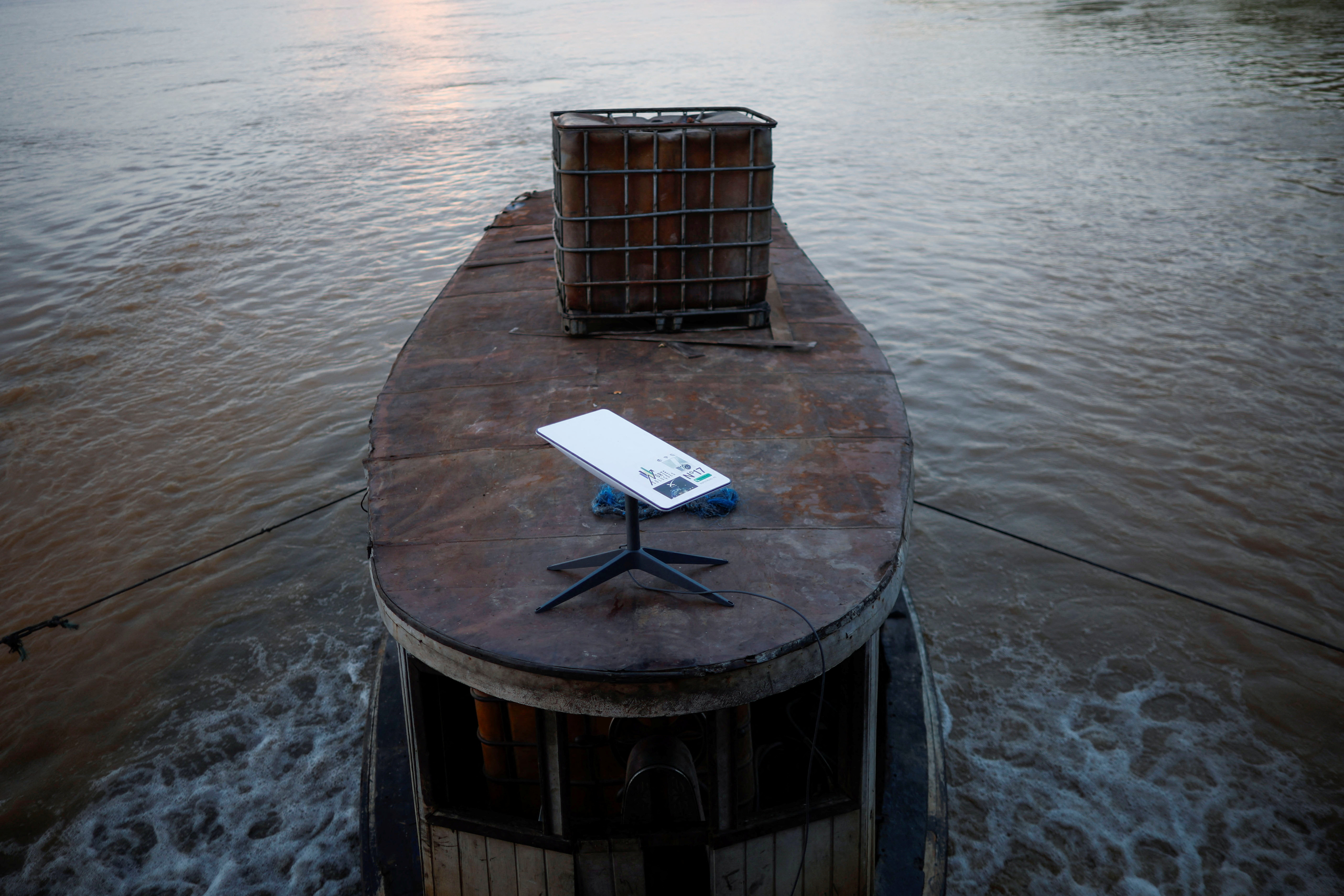 Un barco navegando en un río, con un dispositivo de internet satelital Starlink colocado sobre una mesa en la proa del barco. El agua del río es marrón y el barco lleva un contenedor de gran tamaño en su cubierta. La escena parece capturar un entorno rural o remoto, posiblemente en la región amazónica de Brasil, destacando el uso de tecnología de comunicación avanzada en áreas aisladas.