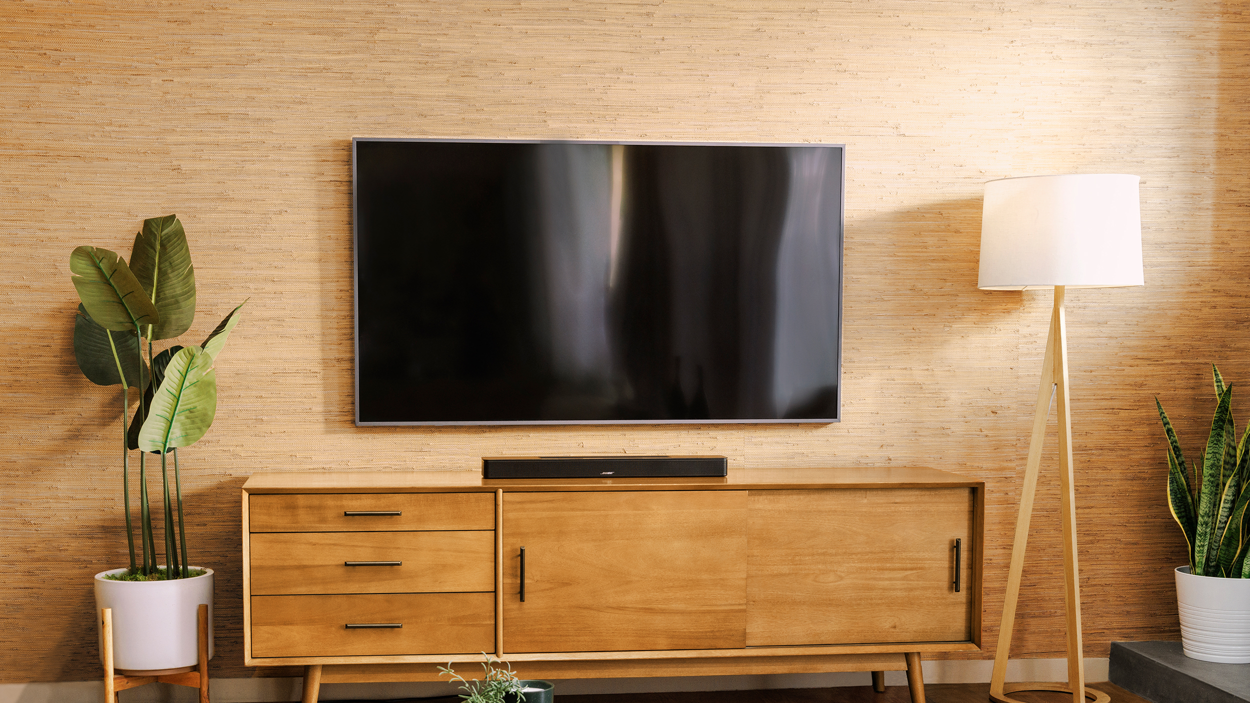 Wide shot of a mounted TV with the Bose Smart Soundbar sitting on an entertainment center below. Potted fern to the left, lamp to the right.