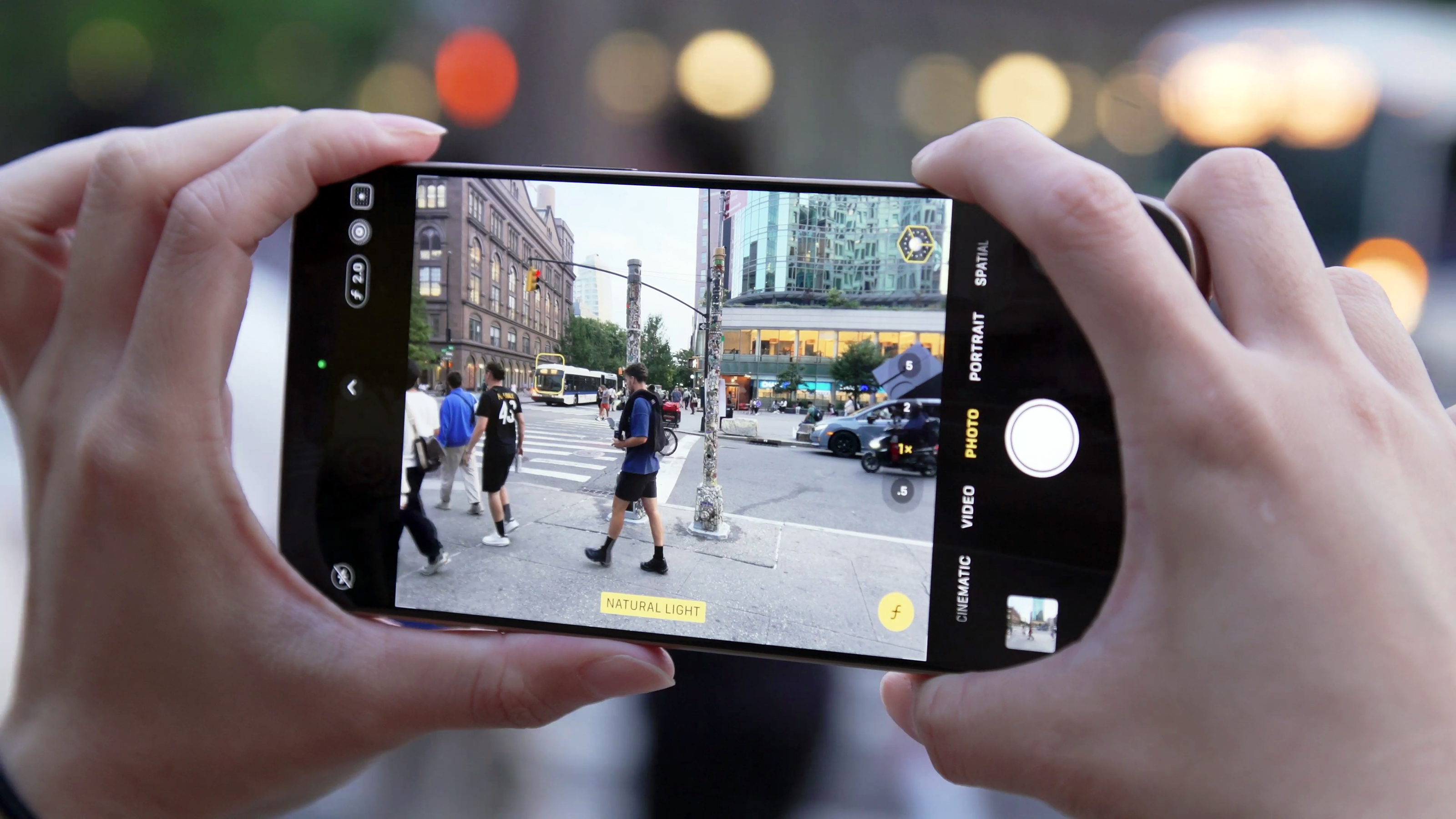 The iPhone 16 Pro held up in mid air by two hands, with the camera app open and showing people walking on a New York City street.