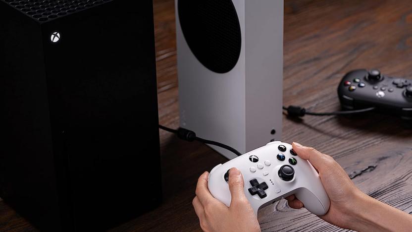 Hands holding a controller with an Xbox on top of a wooden table.