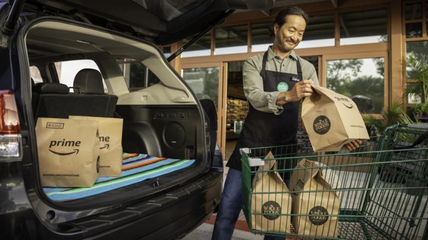 A person loads Amazon Whole Foods grocery bags into a hatchback. They smile as they pick up another bag. Storefront is visible in the background.