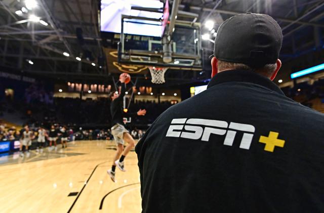A man with an ESPN+ shirt looks at a basketball player on the court making a layup.