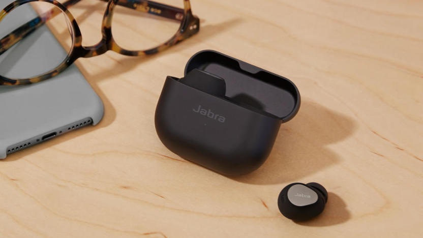 Black wireless earbuds sitting on a desk. One bud is out of the case, sitting on its own, while the case (with the other bud) sits next to it. Glasses and a smartphone are nearby.