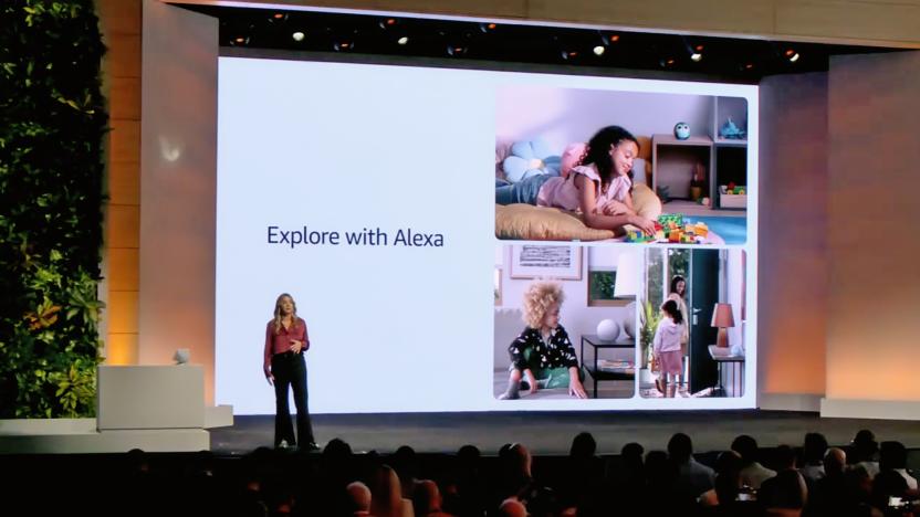 Photo from an Amazon launch event. A presenter stands on stage in front of a huge screen. A slide reads "Explore with Alexa" with a collage of images. Packed audience.