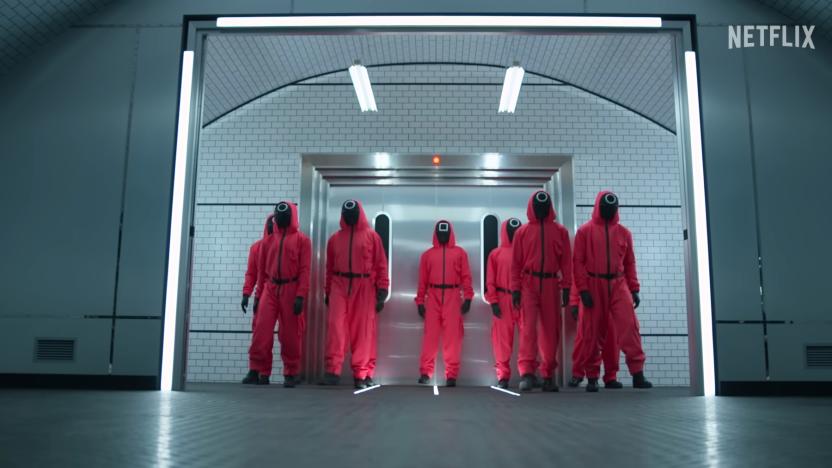 People dressed as Squid Game guards (reddish-pink suits with black faces) stand in a cold industrial building.