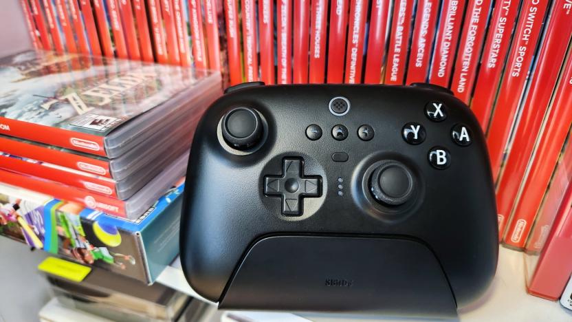 A black video game controller, the 8BitDo Ultimate Bluetooth Controller, rests on a white shelf in front of a row of several red Nintendo Switch video game cases.