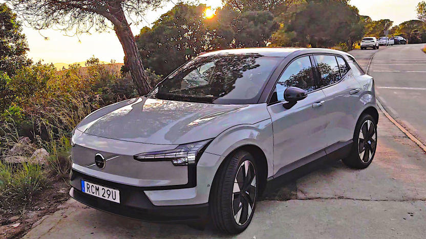 A grey Volvo EX30 is parked at a slight angle on the shoulder of a road with trees, bushes and the sun peeking through the brush in the background.