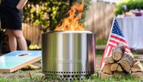 A Solo Stove fire pit sitting on grass next to fire wood and an American flag at a backyard party.