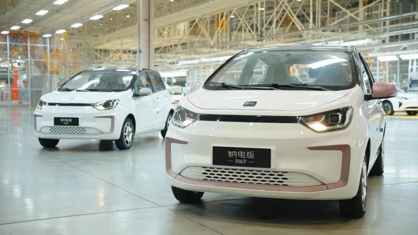 Large car factory or showroom floor featuring two small white electric vehicles. Scaffolding and other cars visible in the background.