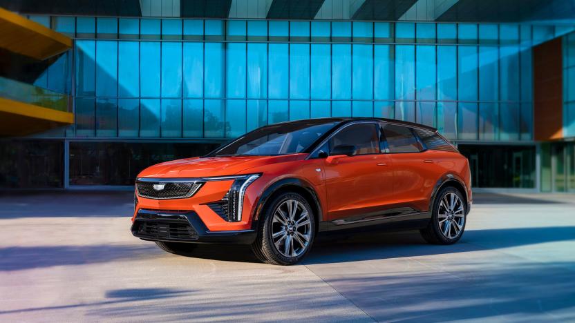 Marketing photo for the Cadillac Optiq. It shows the compact SUV in orange sitting in front of a modern luxury home or garage.