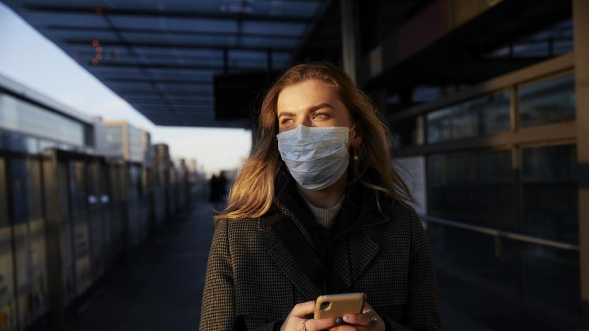Young woman standing on train station wearing protective mask, using phone