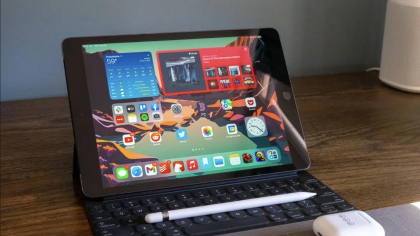 The 9th-gen Apple iPad rests on a brown wooden desk while connected to Apple's Smart Keyboard. A first-gen Apple Pencil rests on top of the keyboard, while a pair of Apple AirPods sit in front of the tablet.