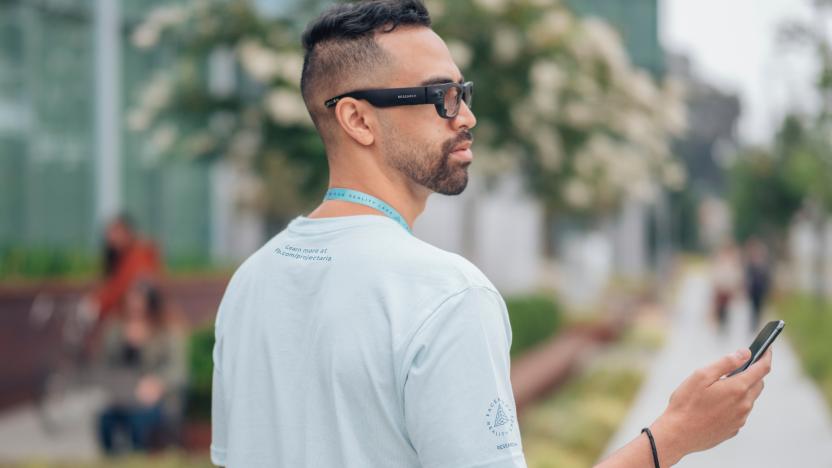 A promo image for the Facebook Project Aria smartglasses, showing a man wearing them outside from his right profile and holding a smartphone.