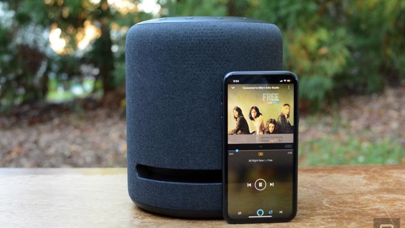 A photo of an Amazon Echo Studio speaker on top of a wooden surface. Propped against it is a phone showing a music streaming app. There are green trees and plants in the background.