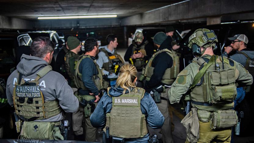 A group of US Marshals Service agents in Operation North Star II gather together in the dark depths of a parking garage with the hood of a car in the foreground.