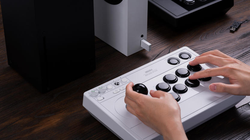 8BitDo marketing photo of a person's hands controlling the company's new arcade stick accessory. The wireless controller has a white body and black sticks / buttons. An Xbox and a spare controller sit on the desk behind it.