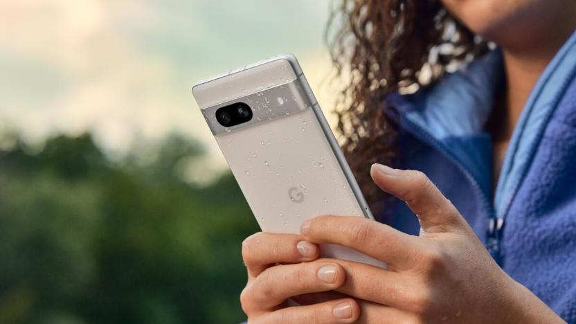 A person holds the snow-colored version of the Google Pixel 7a smartphone. There are water drops on the phone to illustrate its water resistance. 