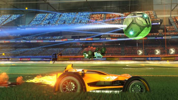 Marketing image from Rocket League, showing a car racing (side view) with a green ball soaring over its head. A truck flips upside down in the background in front of packed stands.
