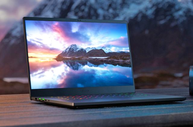 The Razer Blade 15 laptop with 240Hz OLED display seen on a picnic tabletop with mountains in the background and an onscreen image of a larger mountain view with dramatic sky and a reflective lake surface in the foreground.