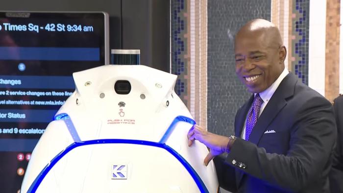 NYC mayor Eric Adams makes one half of a heart sign with his hand against a robot that has no arms.