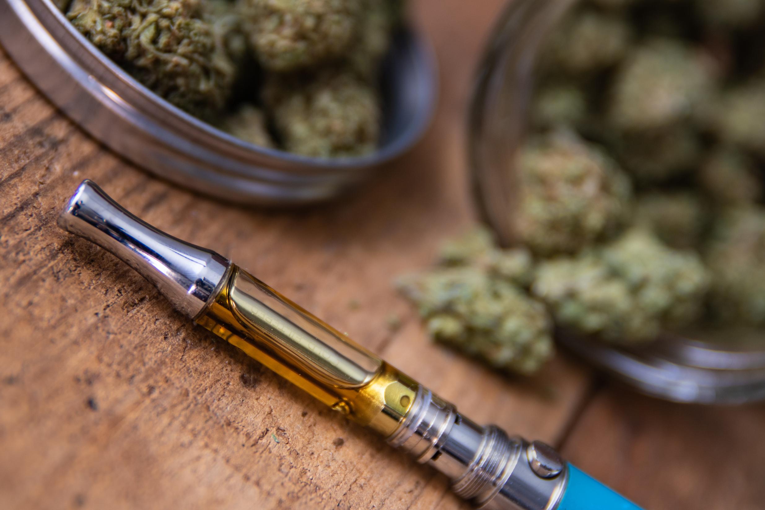 THC oil concentrate filled vape pen on natural wood with an open glass container full of Mango Kush strain marijuana buds grown & sold in dispensaries through-out Southern California.