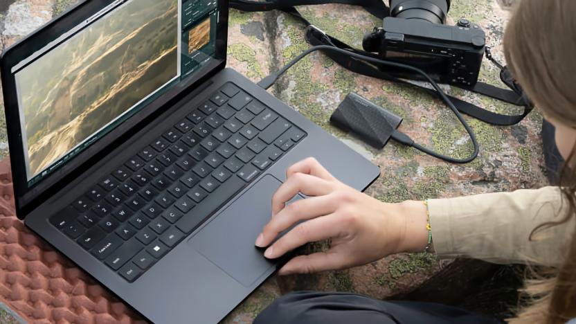 Lifestyle marketing photo of a person using a Windows laptop with a Samsung portable SSD plugged into it. A camera is also nearby, and it all sits on a green and orange surface.