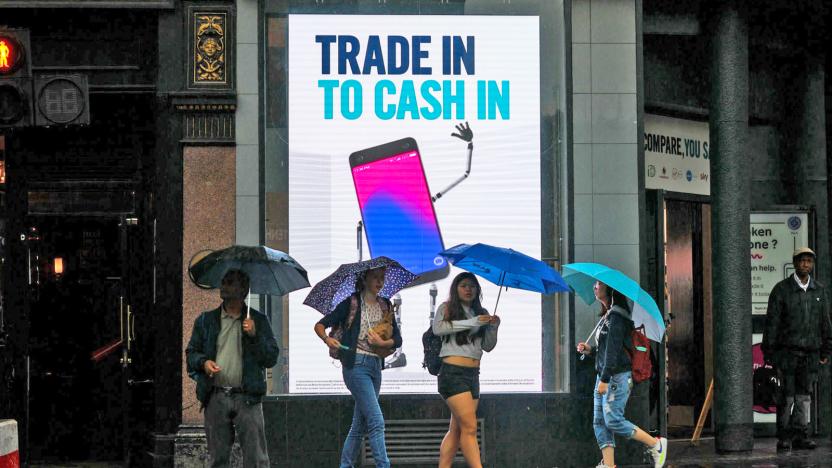 People with umbrellas on a city sidewalk are walking in front of an illuminated sign stating "Trade In to Cash In".