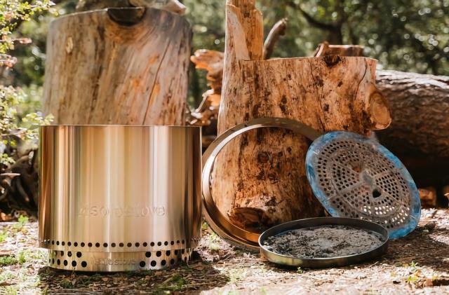 Outdoor scene showing a firepit with cleaning trays leaning against stumps and logs. A lush green foresty scene is in the background.