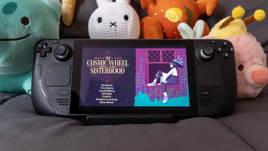 A Steam Deck sitting on a blanket with cute plush characters behind it. On its screen is The Cosmic Wheel Sisterhood’s title screen.