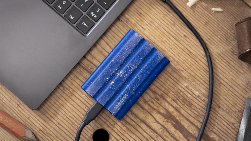 A small and ruggedized portable SSD from Samsung lies on a wooden desk with specs of dust on it, connected to a nearby laptop via USB cable.