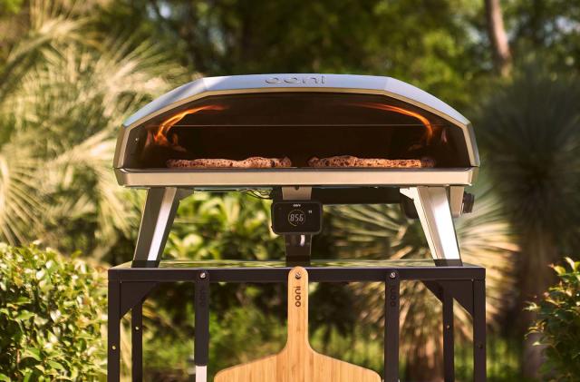 Ooni Koda 2 Max pizza oven sitting on a metal stand in an outdoor setting. Two pizza peels are hanging on the front of the stand and there's visible fire inside the pizza oven.