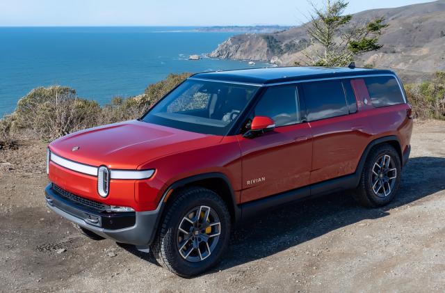 A red Rivian R1S electric SUV parked on a seaside dirt lot.