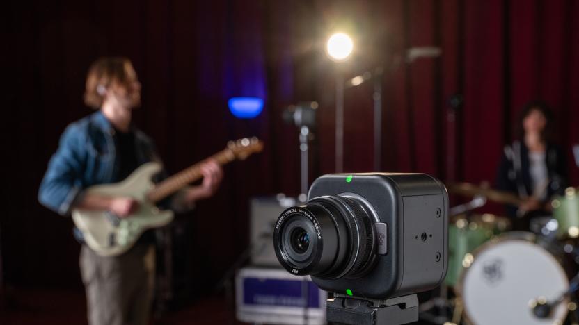 Lifestyle marketing image of the Logitech Mevo Core livestreaming camera. It sits on a tripod with a rock band playing in a studio behind them.